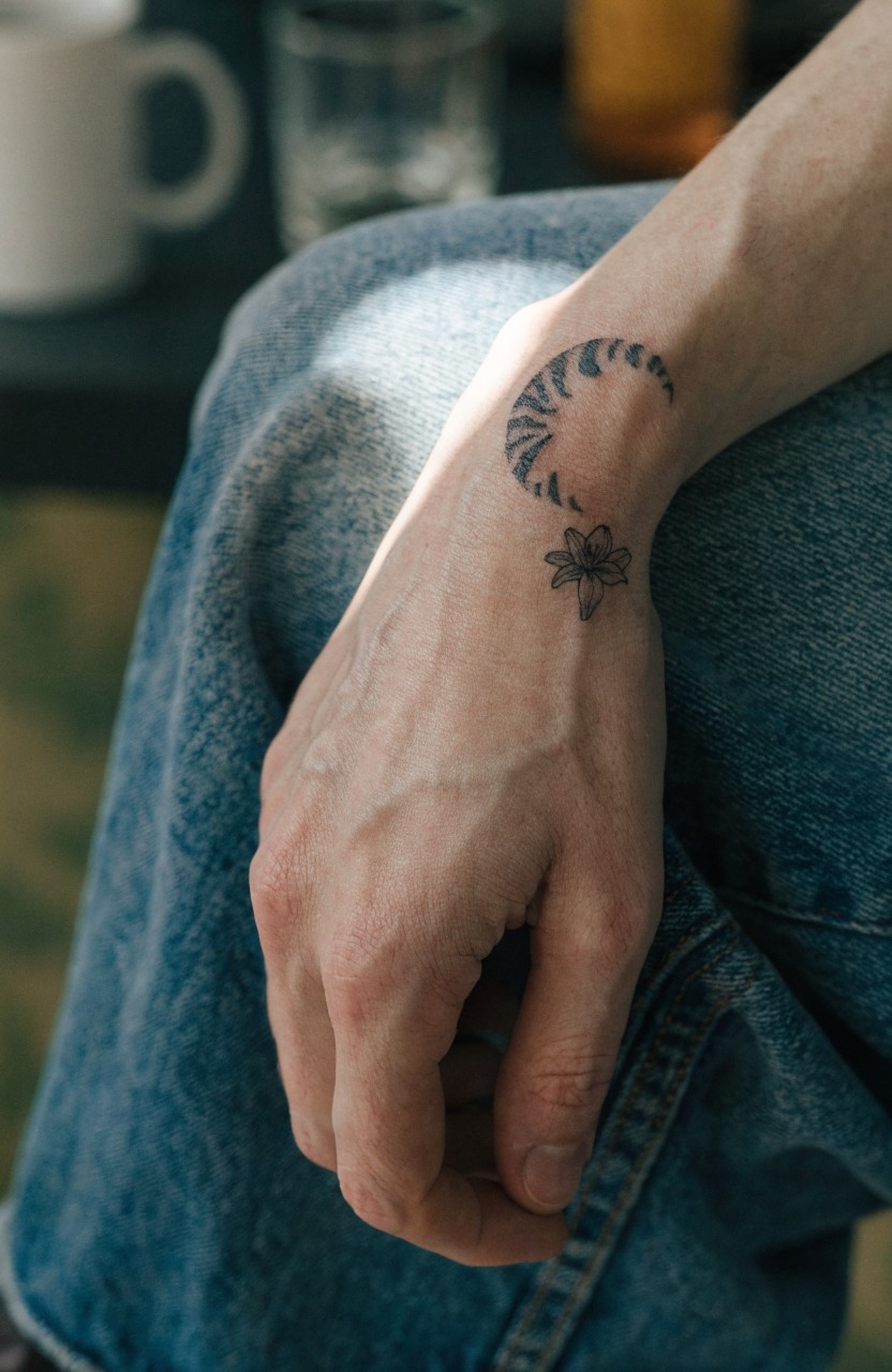 Minimalist black line tattoo of tiger-striped crescent moon with small flower on inner wrist