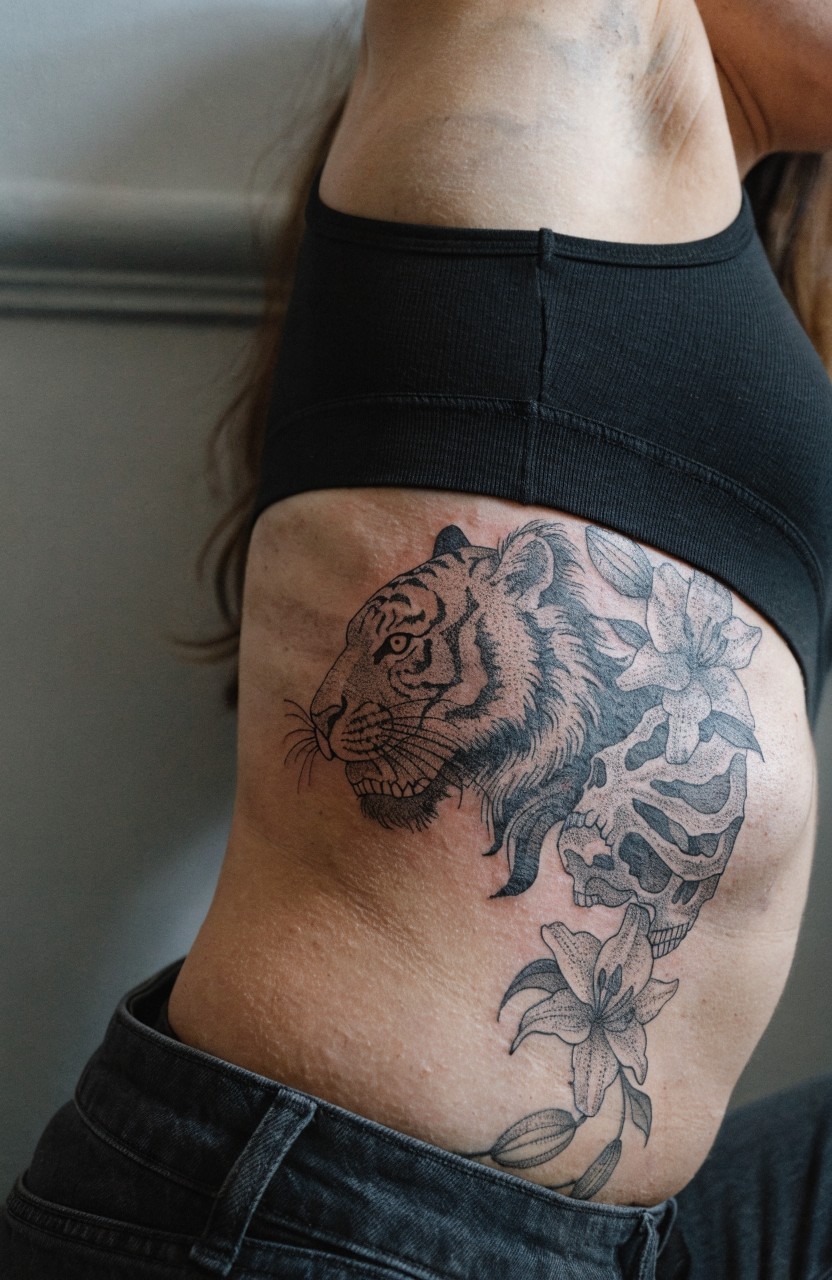 Black and gray realistic tiger head tattoo with lilies and skull on a woman's side torso and ribs