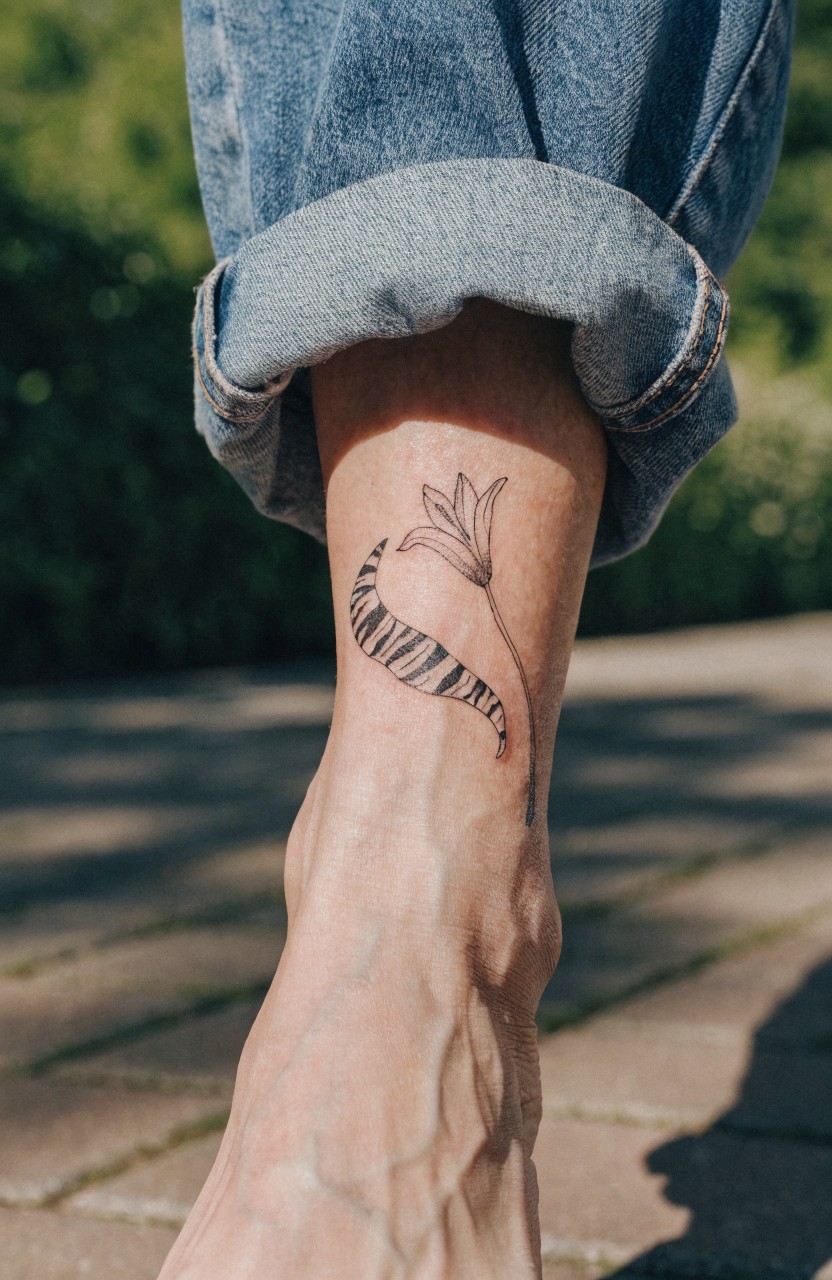 Fine line black ink tattoo of a lily flower with zebra stripes curving around a man's ankle on tanned skin, jeans rolled up outdoors