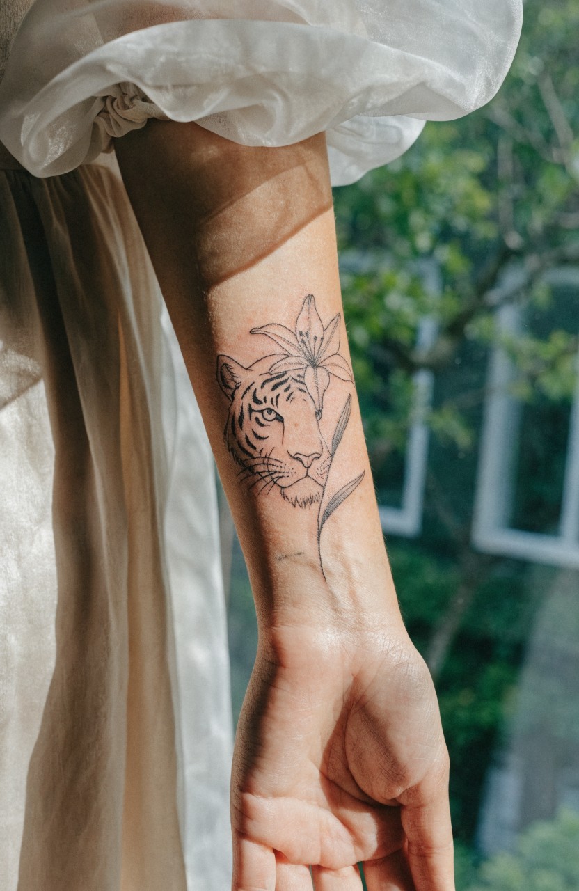 Line art tattoo of tiger head with lily flower on inner forearm, thin black outlines on pale skin