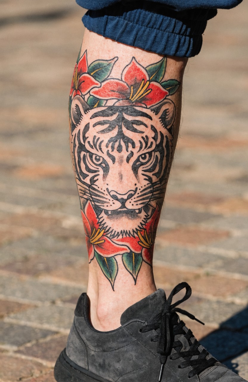 Black and white tiger head tattoo on a calf surrounded by red lilies, viewed from the front with sweatpants above and sneakers below