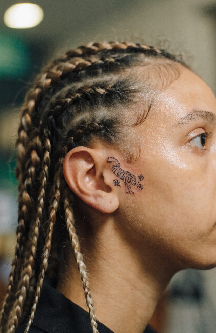 Small fine line black ink tiger face with flowers tattoo near the ear and cheek on a person with braided hair