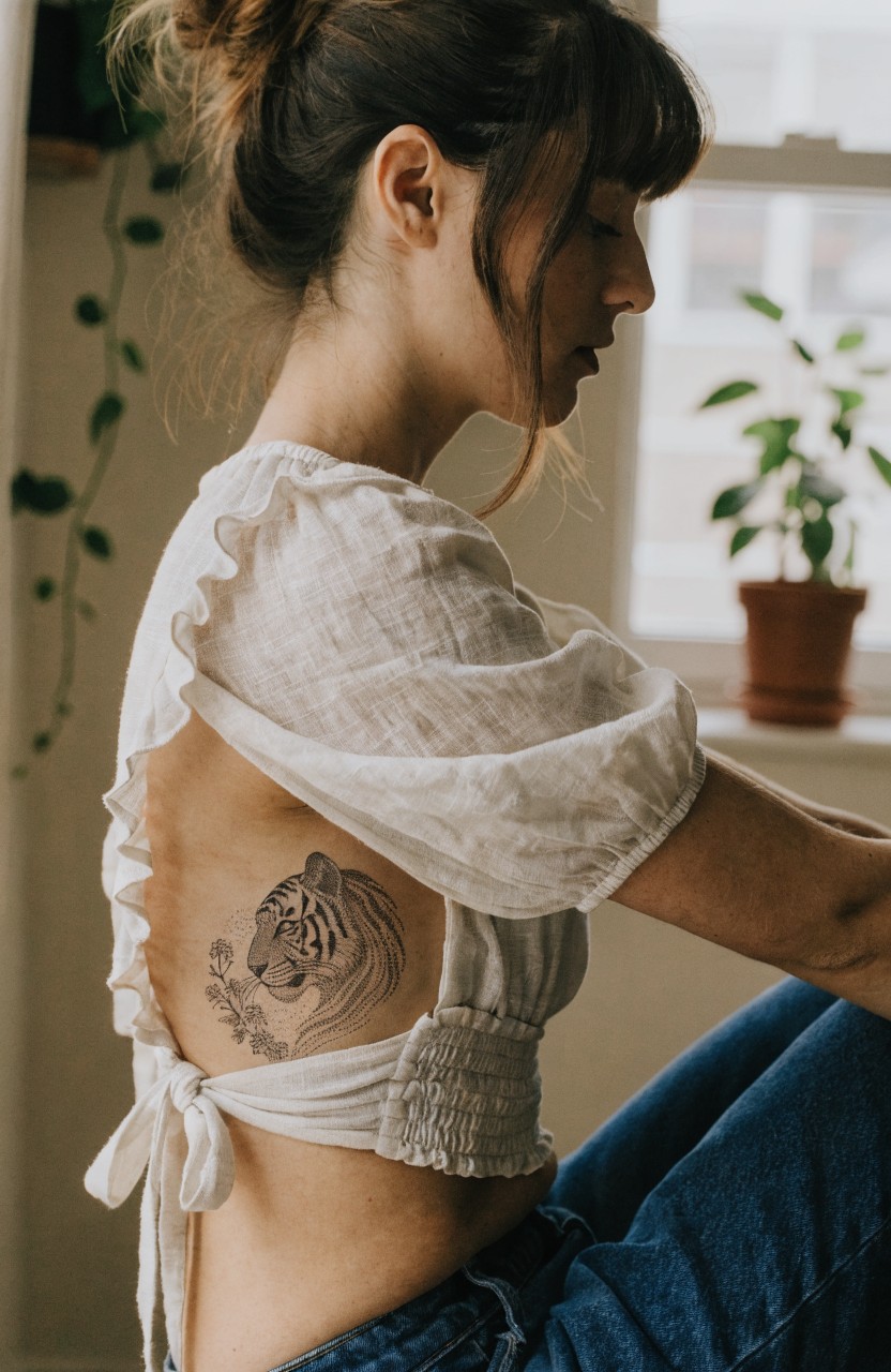 Detailed black and gray tiger face tattoo with integrated flowers on a woman's lower back and side, shown from a profile view