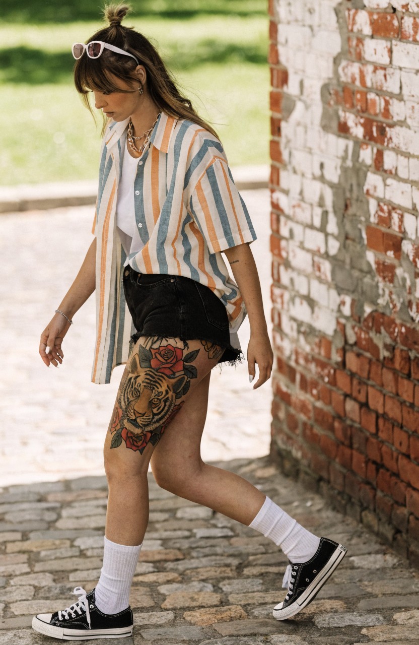 Woman modeling a large colorful tiger face with red roses tattoo on her upper thigh, wearing striped shirt, black shorts, white socks and black sneakers against brick wall