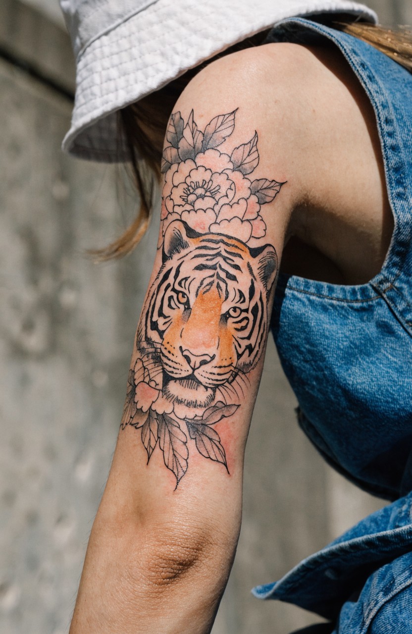 Woman's upper arm tattoo of a detailed tiger head in black linework and orange shading, framed by large peony flowers in fine black ink