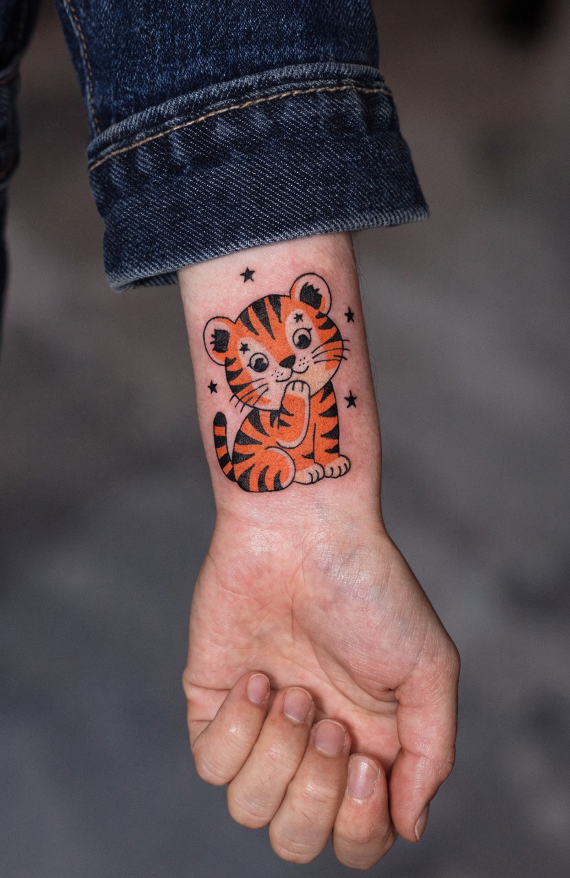 Colorful cartoon tiger cub tattoo on inner wrist with orange fur, black stripes, shy paw-to-mouth pose, and sparkling stars around it