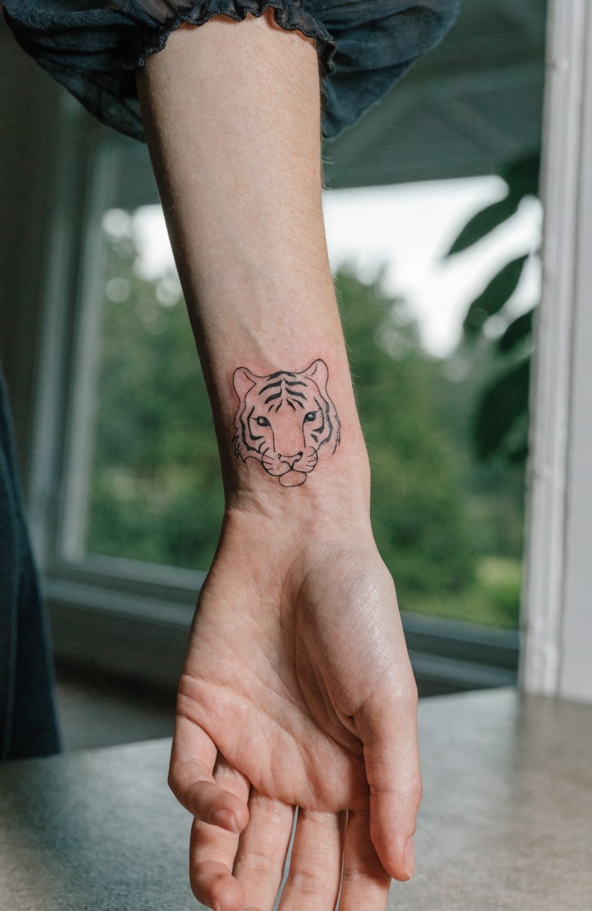 Small black linework tiger head tattoo on woman's inner wrist with subtle shading