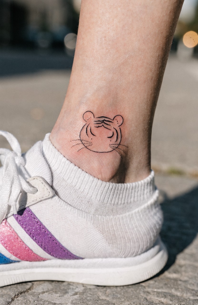 Minimalist black line art tiger face tattoo on a woman's ankle above the bone, next to pulled down white crew sock and pink striped sneakers on city pavement