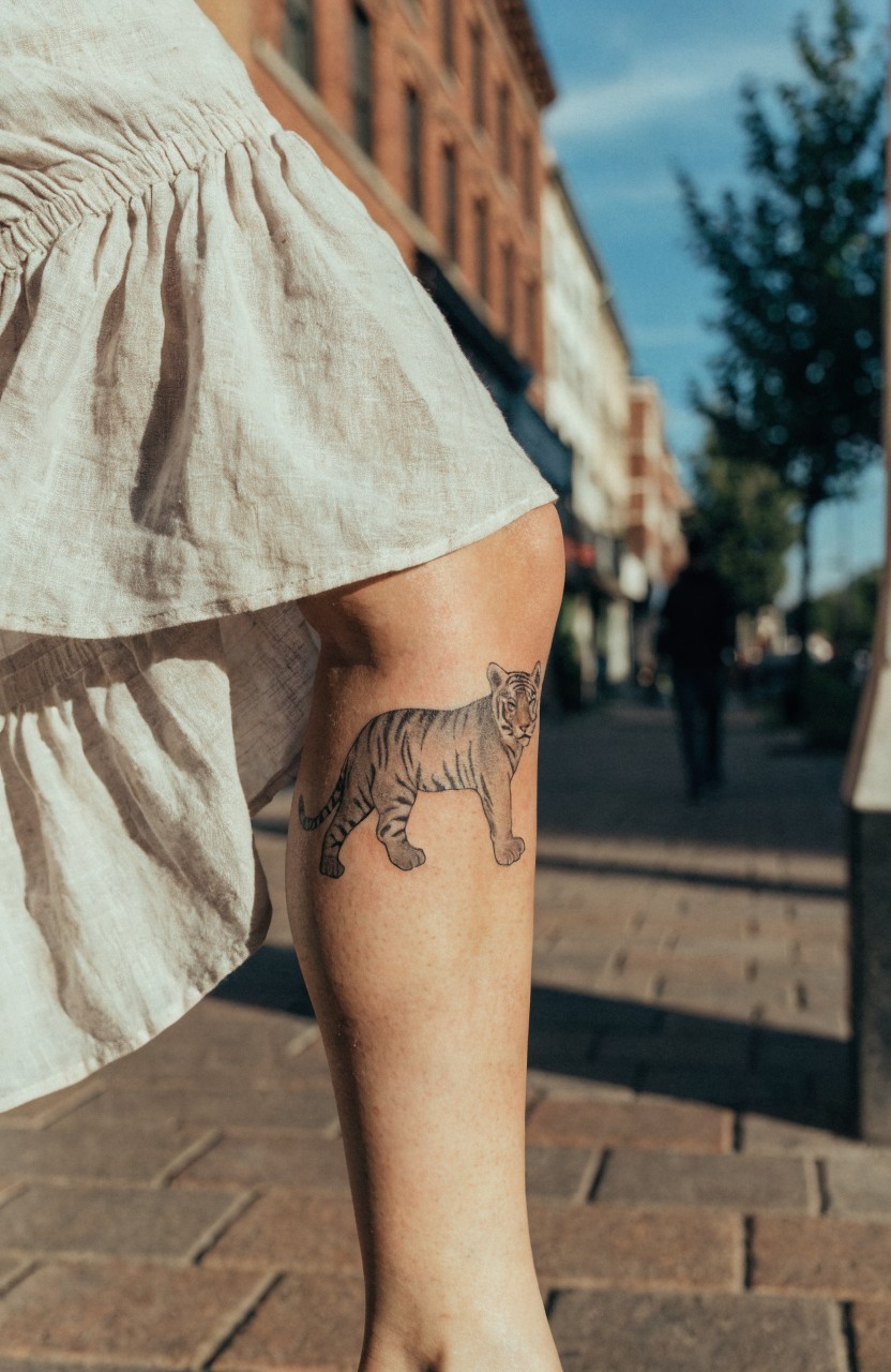 Black and gray tiger tattoo in walking pose on woman's lower leg, shown outdoors on paved street with brick buildings and trees in background