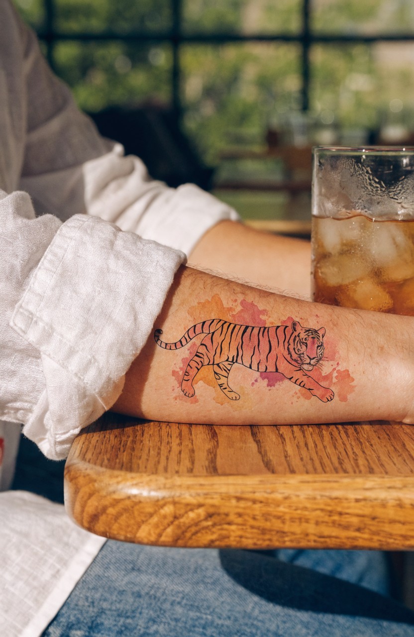 Watercolor tiger tattoo in pink and orange with splash effects on a woman's outer forearm next to a glass of iced tea