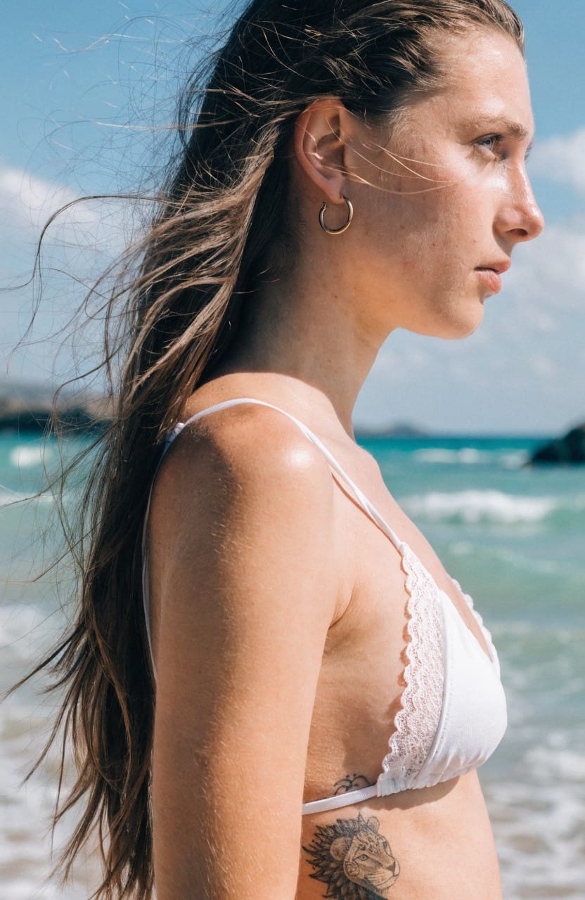 Close-up of a woman's side torso revealing a realistic black and grey lion head tattoo under a white lace bralette at the beach