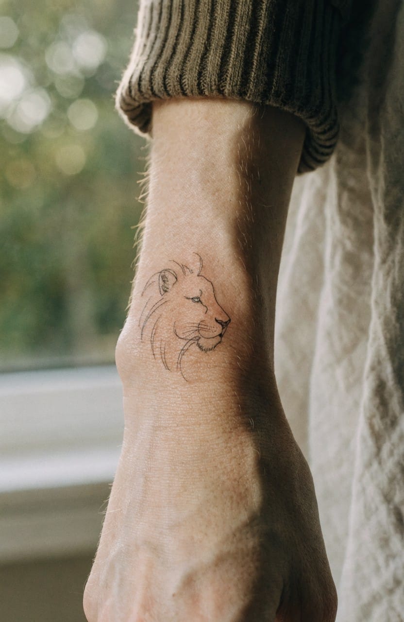 Minimalist fine line lion head tattoo in profile on a woman's inner forearm, black ink with light shading, viewed against a window backdrop