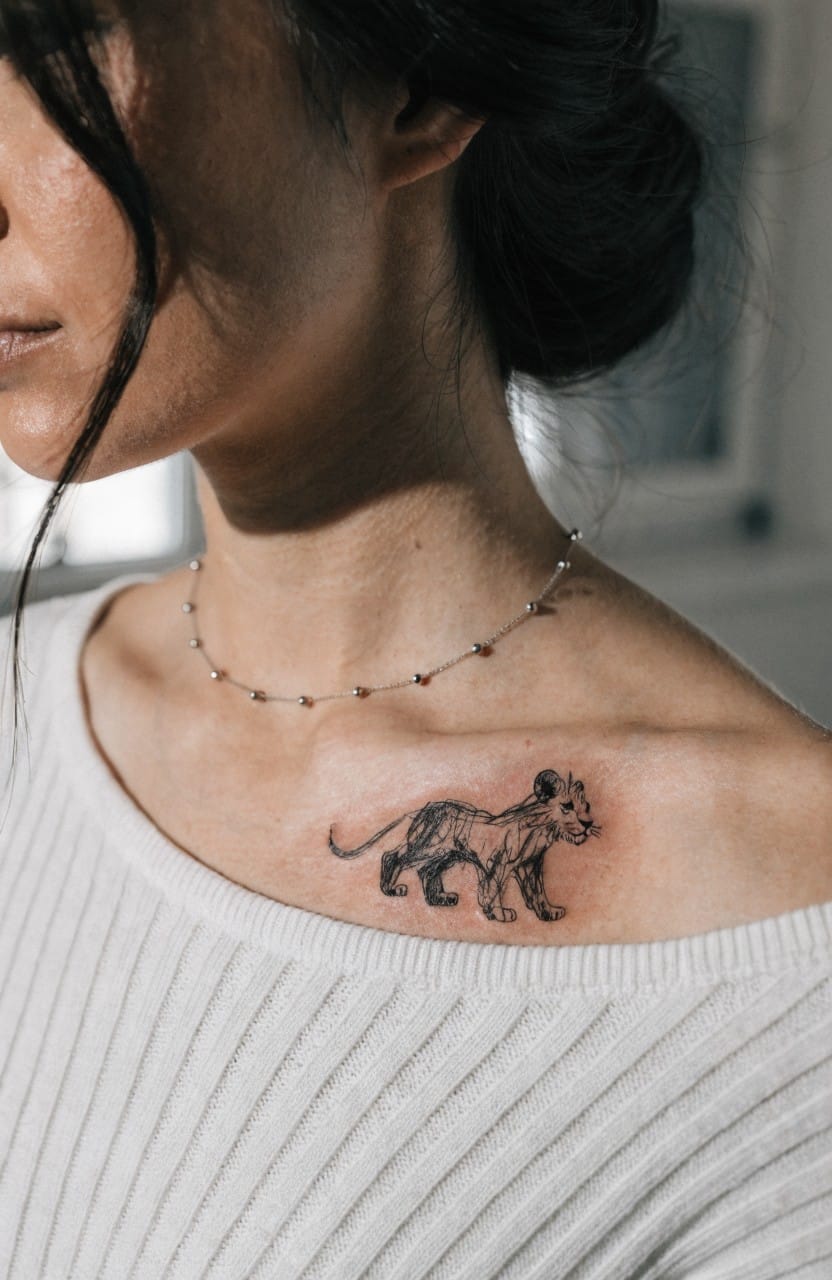 Black line art tattoo of a young lion cub on a woman's shoulder and collarbone, fine lines with subtle shading