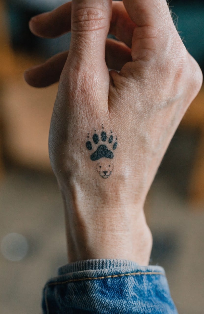 Minimalist black ink lion paw print tattoo on the back of a hand with a small lion face in the center pad and dot accents