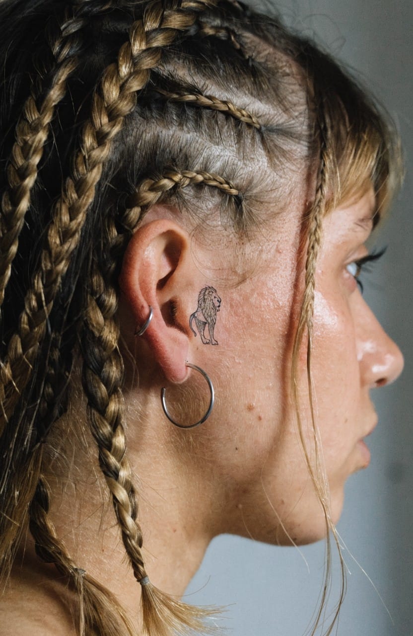 Side profile close-up of a woman with blonde cornrows and hoop earrings displaying a small minimalist black lion head tattoo on her cheek near the ear