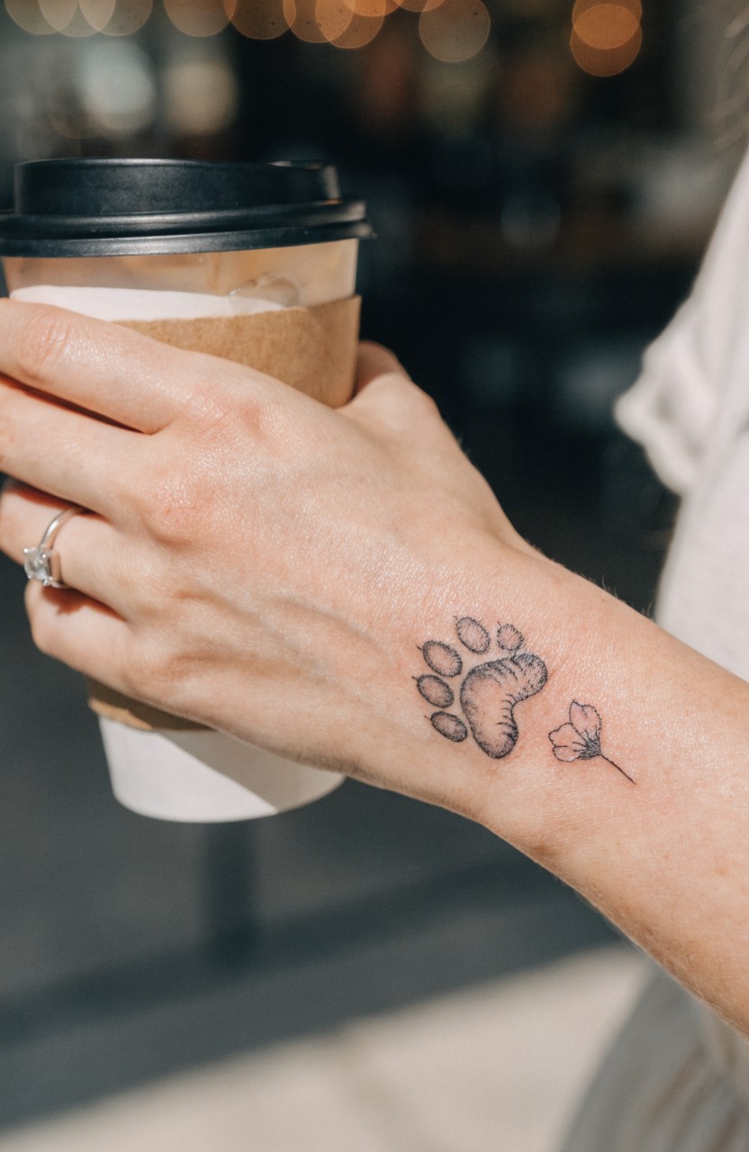 Minimalist black line tattoo of a paw print with small flower on a woman's inner wrist, hand holding a paper coffee cup against a blurred cafe background