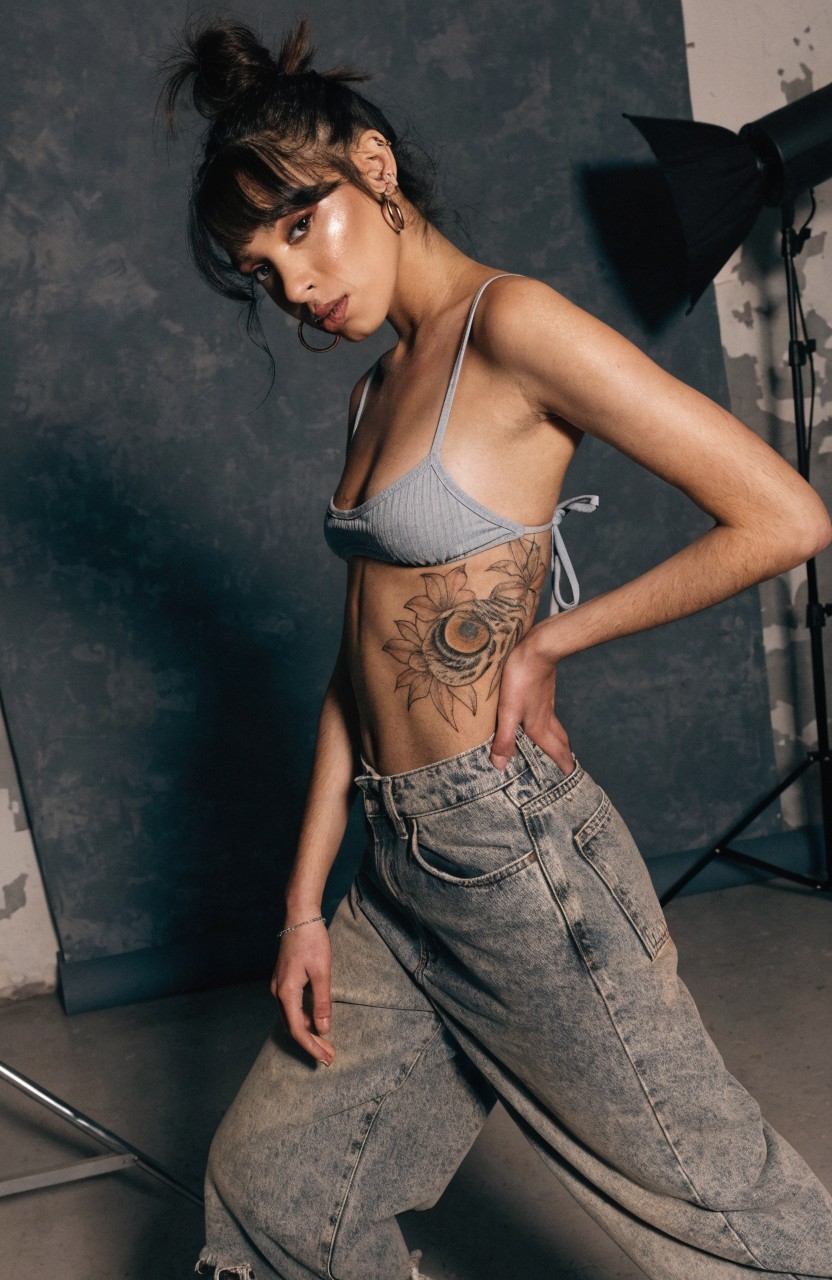 Model showing large black and grey realistic sunflower tattoo on side ribs and torso, posed in studio with light blue bralette and baggy jeans