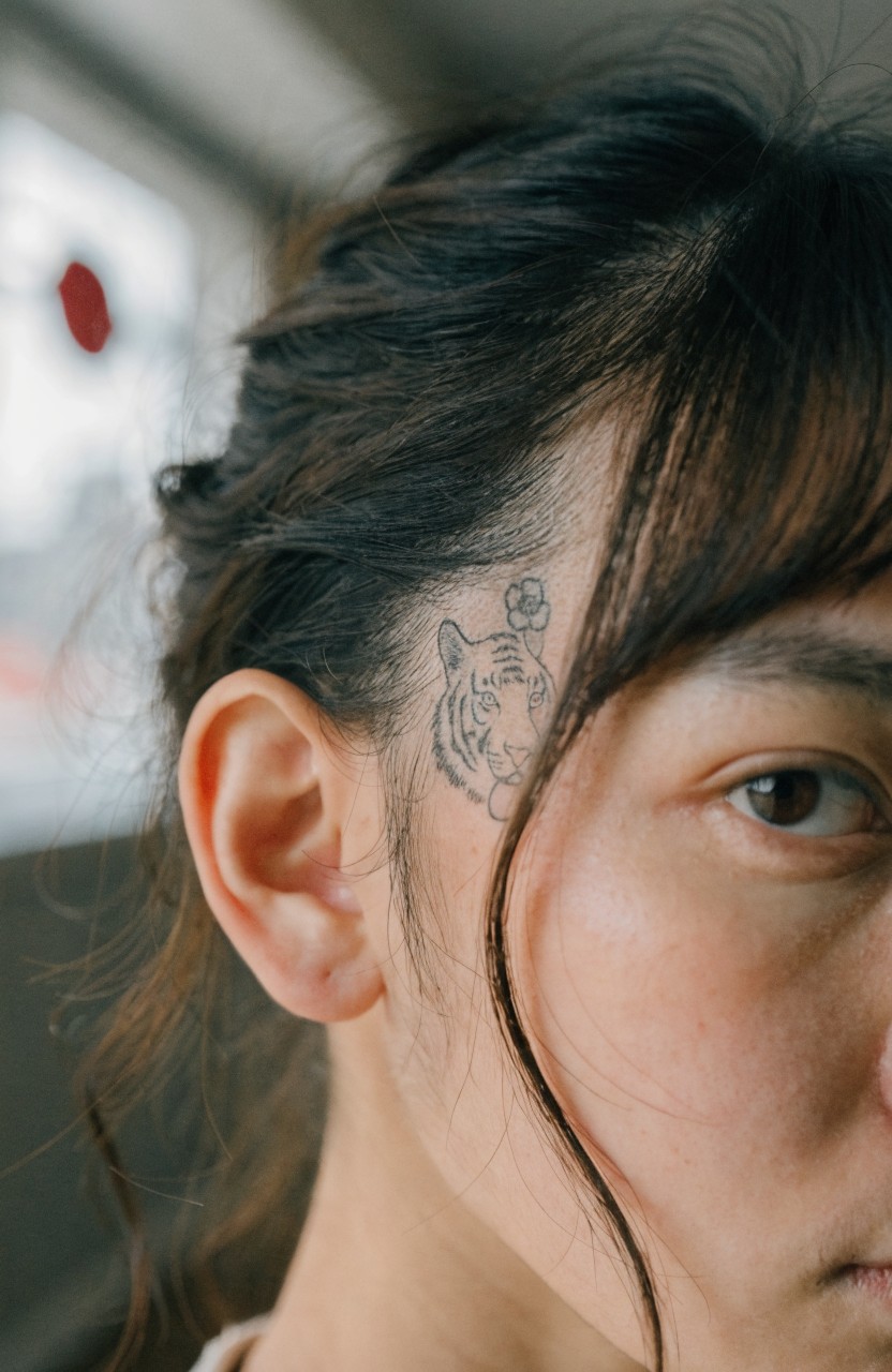 Fine line black ink tattoo of a tiger head and flower on the temple and behind the ear of a woman with dark hair