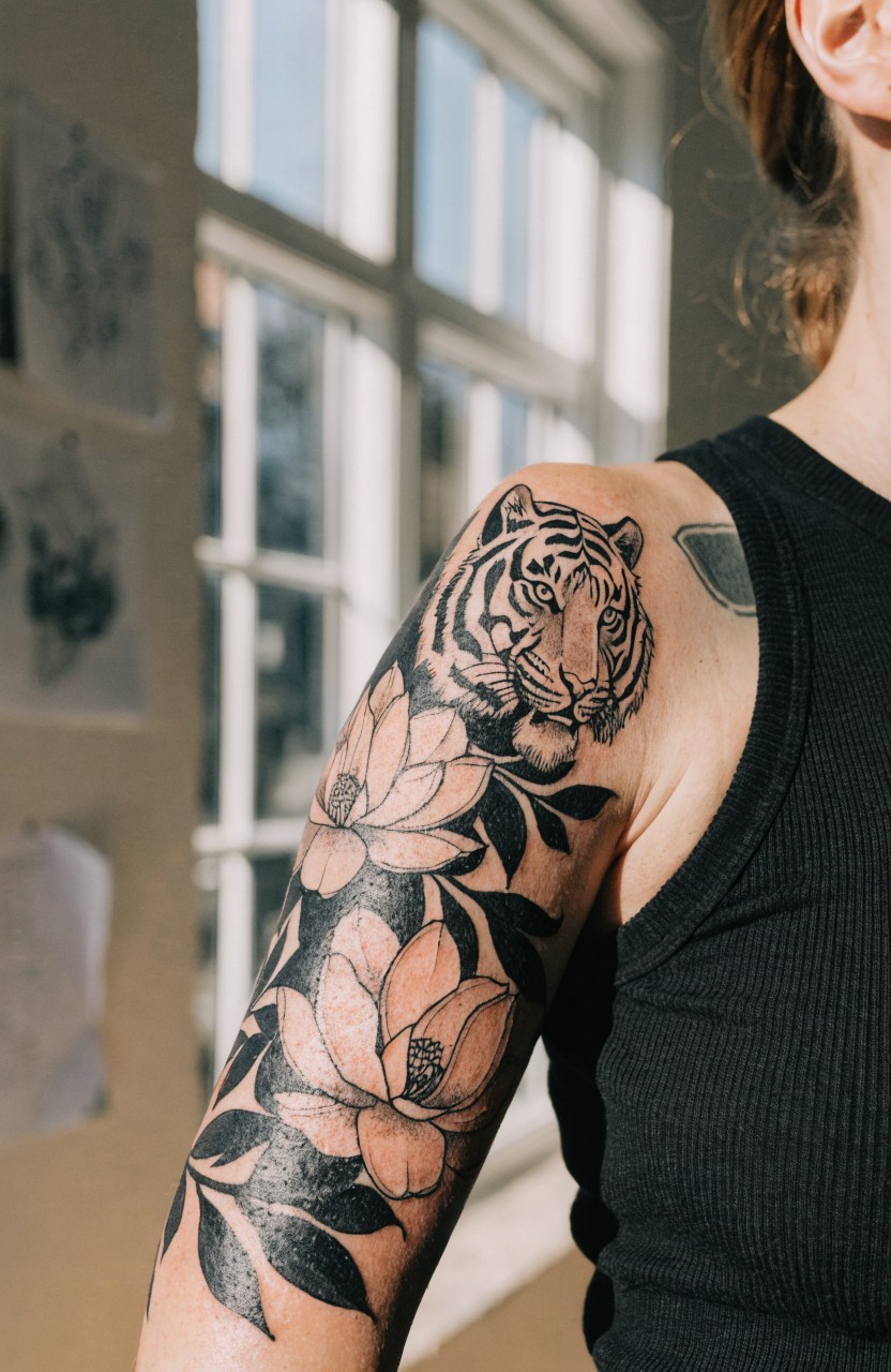Black linework tattoo of tiger head on woman's upper arm framed by magnolia flowers and leaves