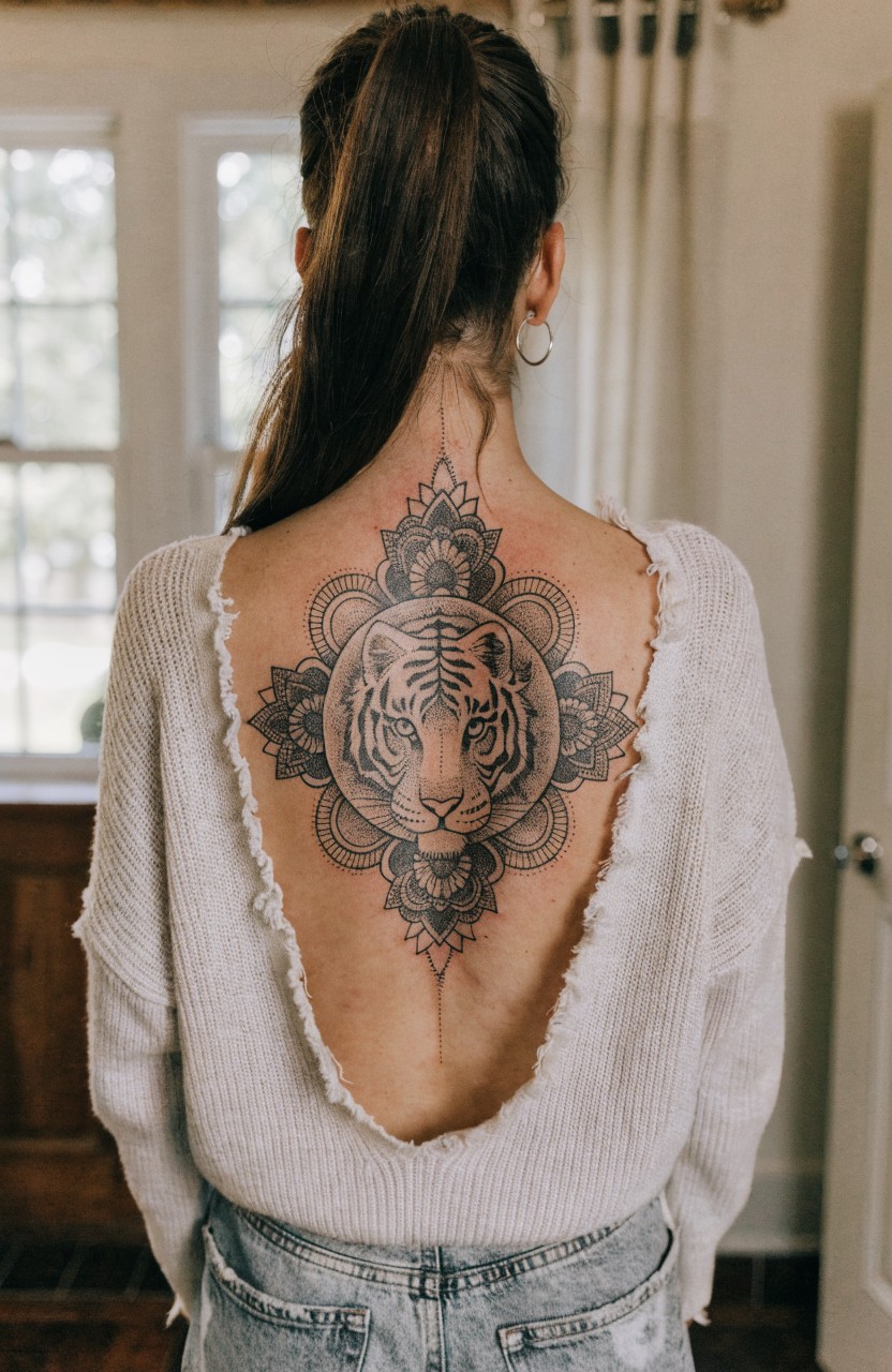 Large black ink mandala tattoo with central tiger head on woman's upper back, seen through backless white sweater