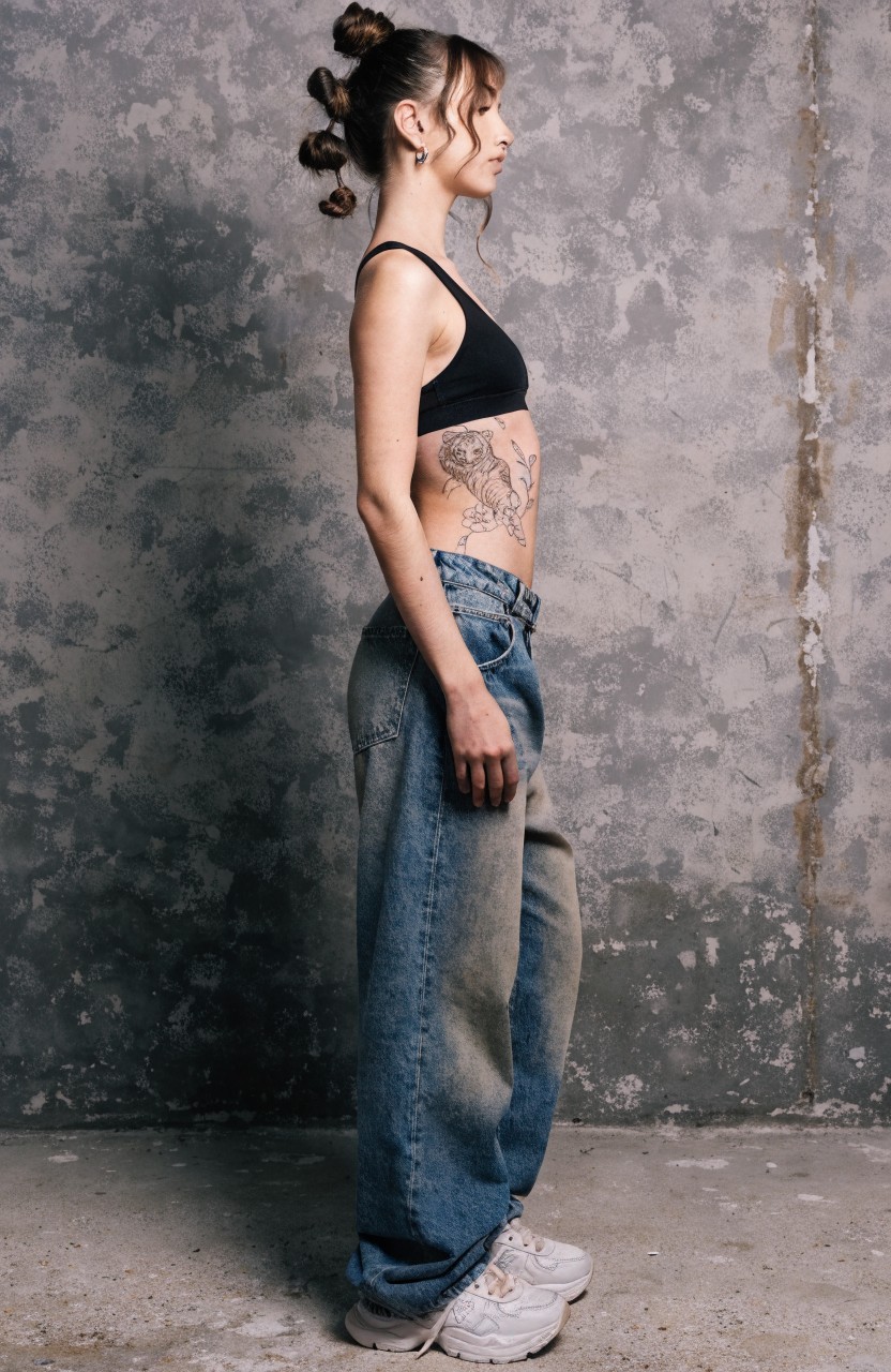 Woman in side profile with large realistic black ink tiger tattoo on her side torso, wearing a black crop top, baggy jeans, and white sneakers against a textured wall