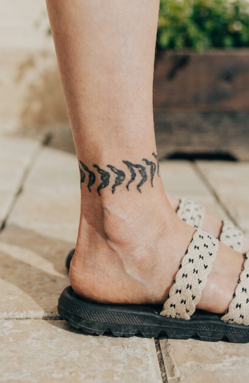 Black linework tattoo of abstract wavy tiger stripes forming an anklet around a woman's ankle, above white braided sandals on stone tiles