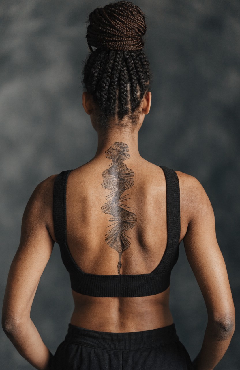 Black ink floral tattoo centered on a woman's upper back and neck following the spine with blooming flower at top and twisting stem below