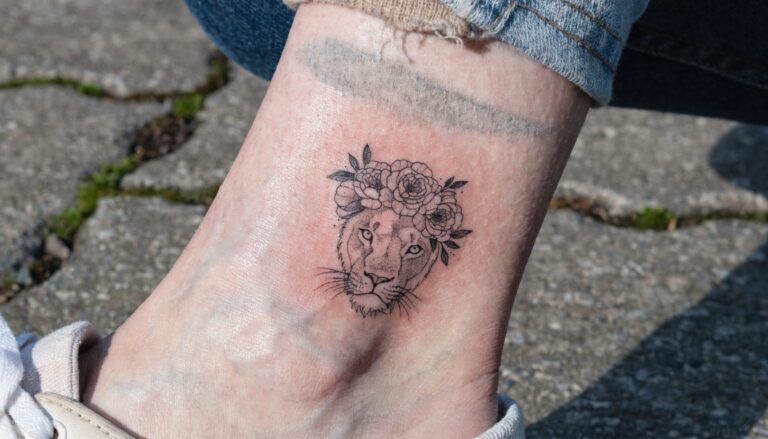 21 Creative Lion and Flower Tattoo Ideas With a Feminine Twist Black linework tattoo of lioness head crowned with peonies on ankle bone, shown on leg with white sneakers pulled up over socks on cobblestone path