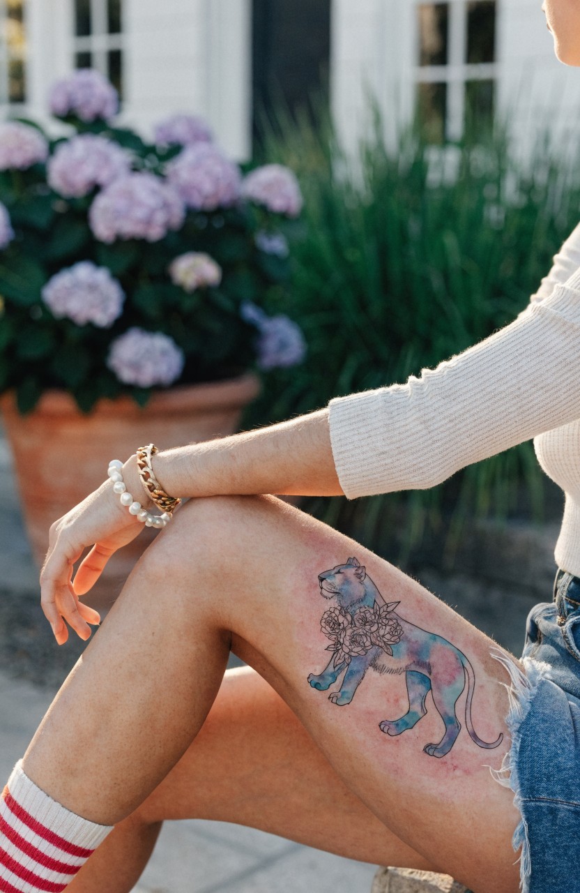 Colorful watercolor lion tattoo with integrated flowers on outer thigh, in shades of blue and purple