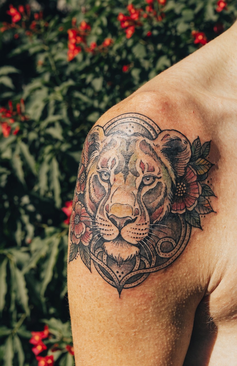 Shoulder tattoo of a detailed lioness head in black and gray shading framed by red flowers and ornate borders on bare skin amid green plants