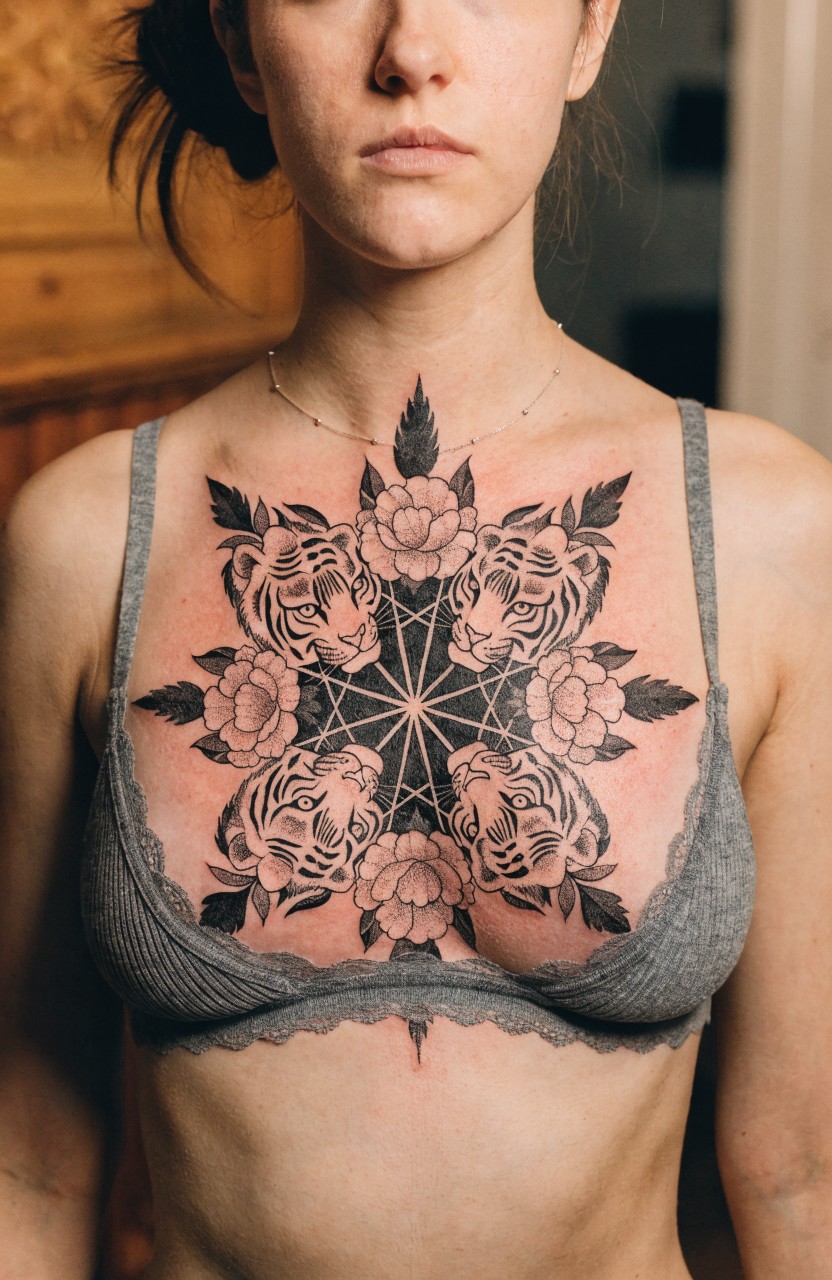 Woman's chest tattooed with a large symmetrical black ink mandala of tiger faces surrounded by flowers and leaves centered on the sternum