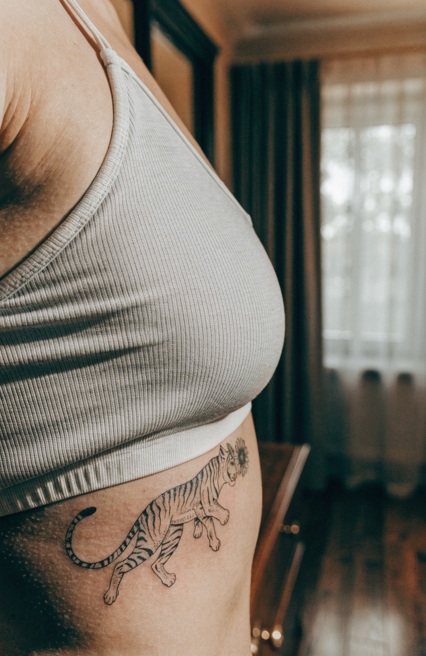 Realistic black and gray tiger tattoo curving along a woman's side ribs under a gray tank top