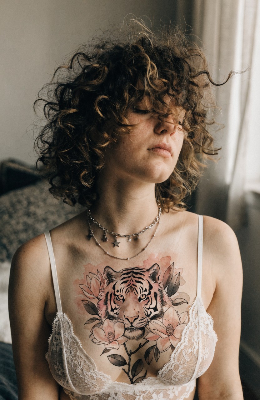 Closeup of a woman with curly brown hair and a large colorful tiger face tattoo framed by pink flowers on her upper chest above a white lace bra