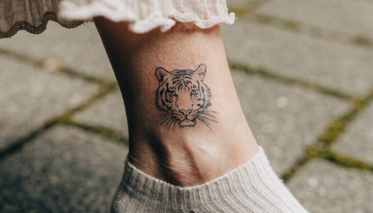 Black linework tiger face tattoo on ankle bone above white sock on paved path
