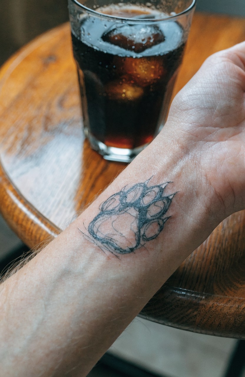 Sketchy black ink paw print tattoo on inner wrist beside cola glass on wood table