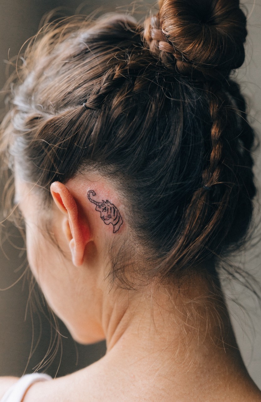 Close-up side profile of a woman with long brown hair in a bun revealing a small black linework tiger head tattoo behind her ear