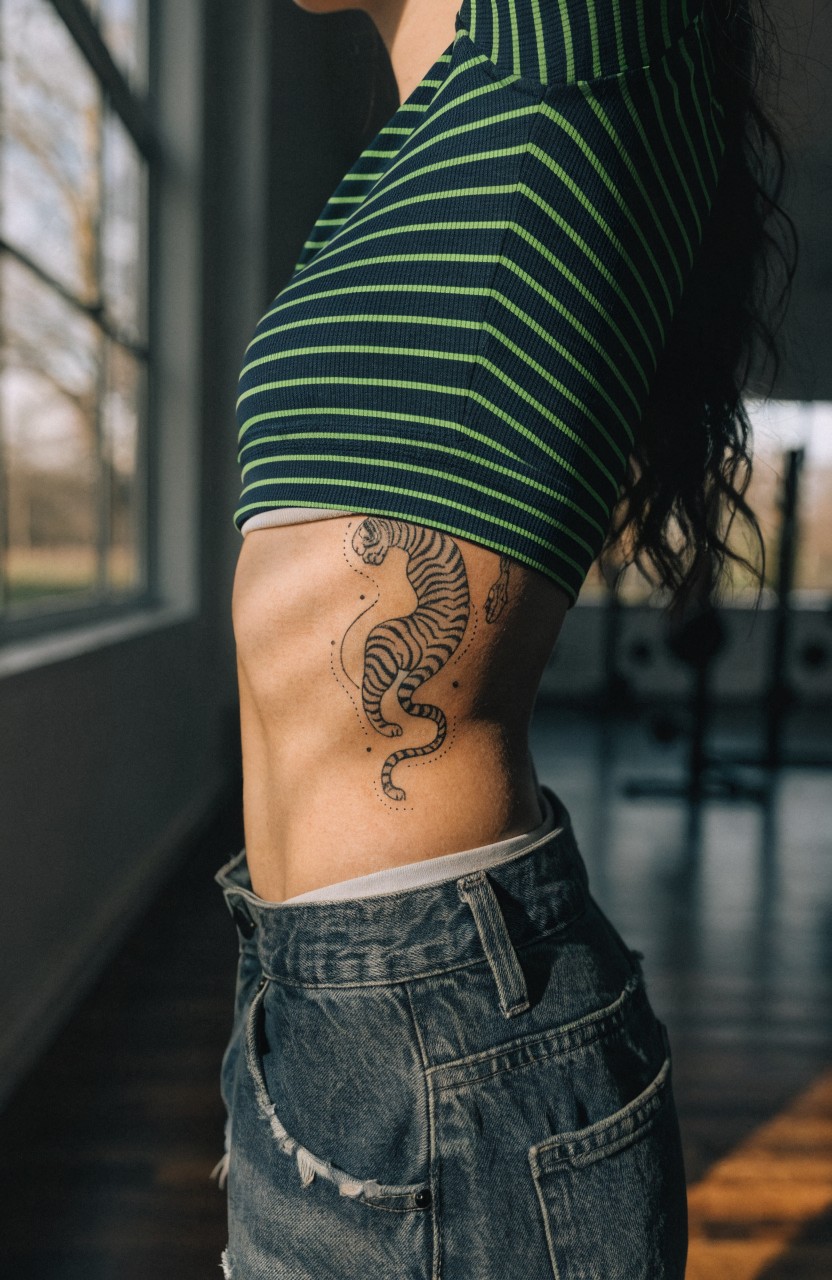 Black linework tiger tattoo curving from ribs to hip along a woman's side torso