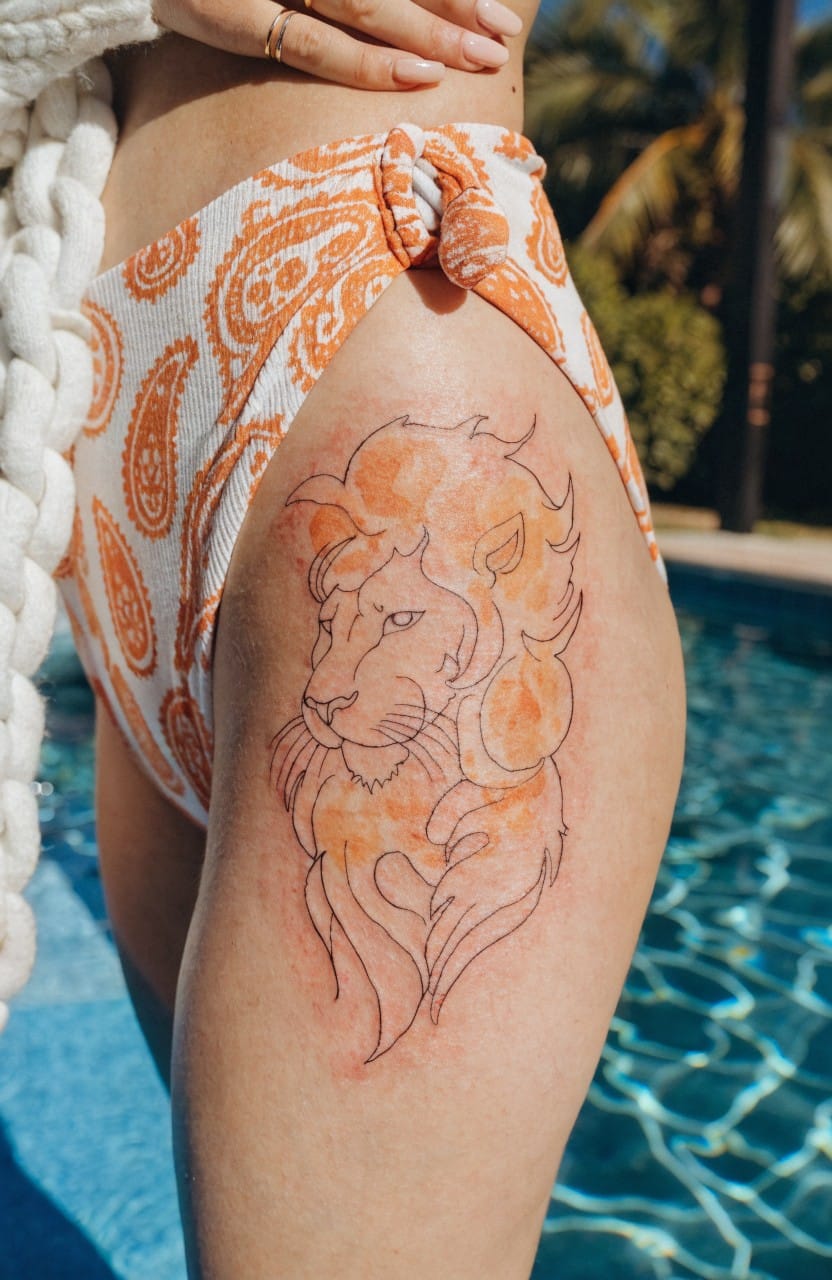Fine line tattoo of a lion head in profile on a woman's upper thigh with orange watercolor shading on the mane