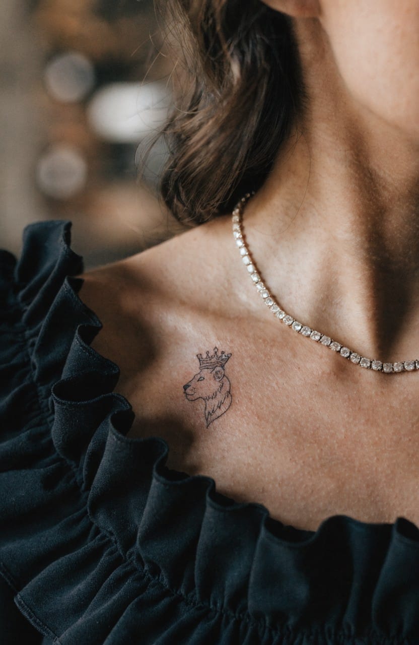 Woman's shoulder with small black linework lion head and crown tattoo