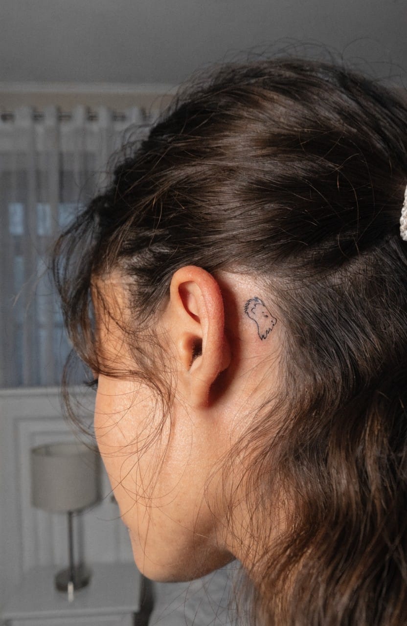 Woman's profile view with small black outline lion head tattoo behind her ear under wavy brown hair in a bedroom setting