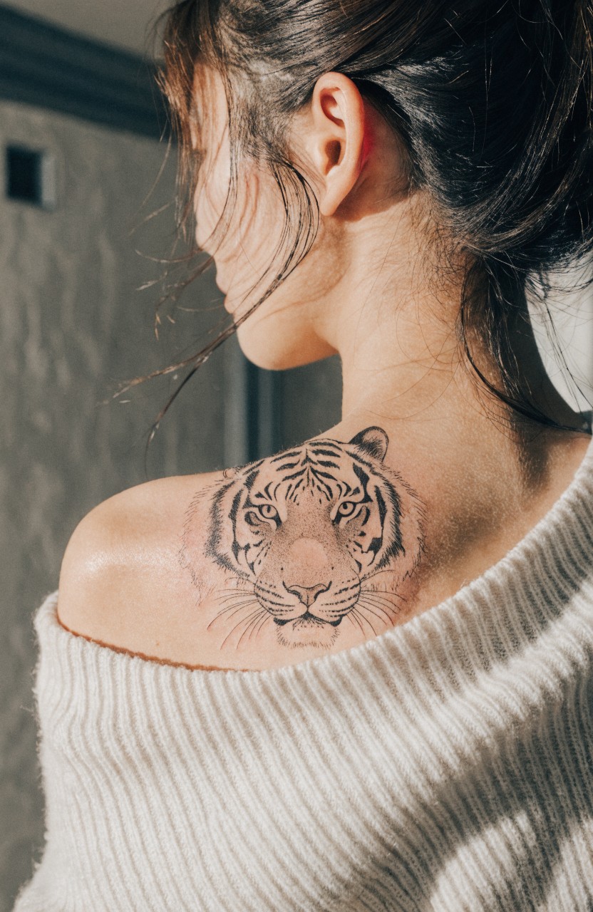 Black ink tiger head tattoo on a woman's shoulder blade, viewed from behind with off-shoulder white sweater and hair pulled aside