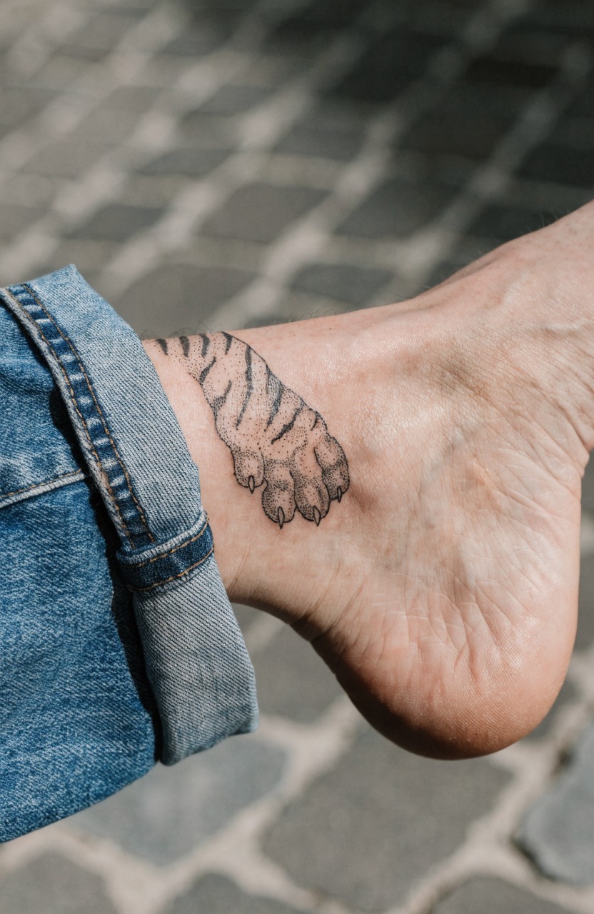Black ink tiger paw tattoo on outer ankle with shading on stripes and claws, woman's foot in rolled jeans on tiled ground