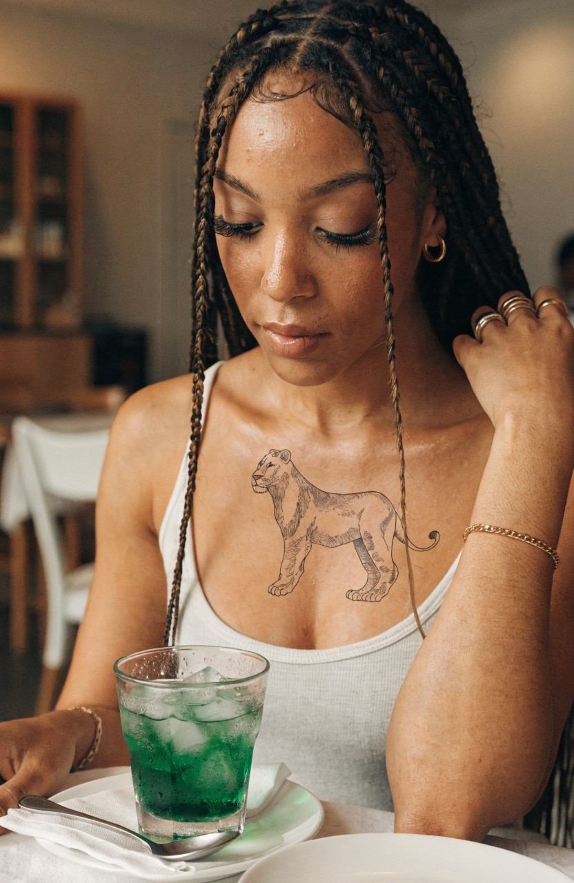Black linework lion tattoo on a woman's upper chest over her right breast, detailed outline with light shading, woman with braids holding a green drink at a table