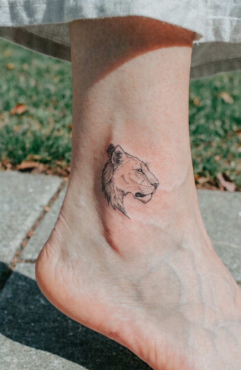 Small fine line lioness head tattoo on a woman's outer ankle in black ink with light shading