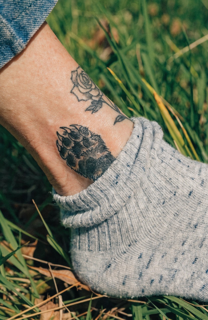 Black and grey rose and shaded paw print tattoo on a woman's ankle over grass, with cuffed jeans and socks