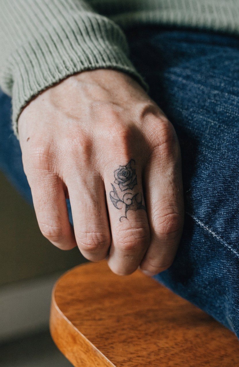 Close-up of a small black ink rose tattoo on ring finger, hand in green sweater and jeans on wooden chair