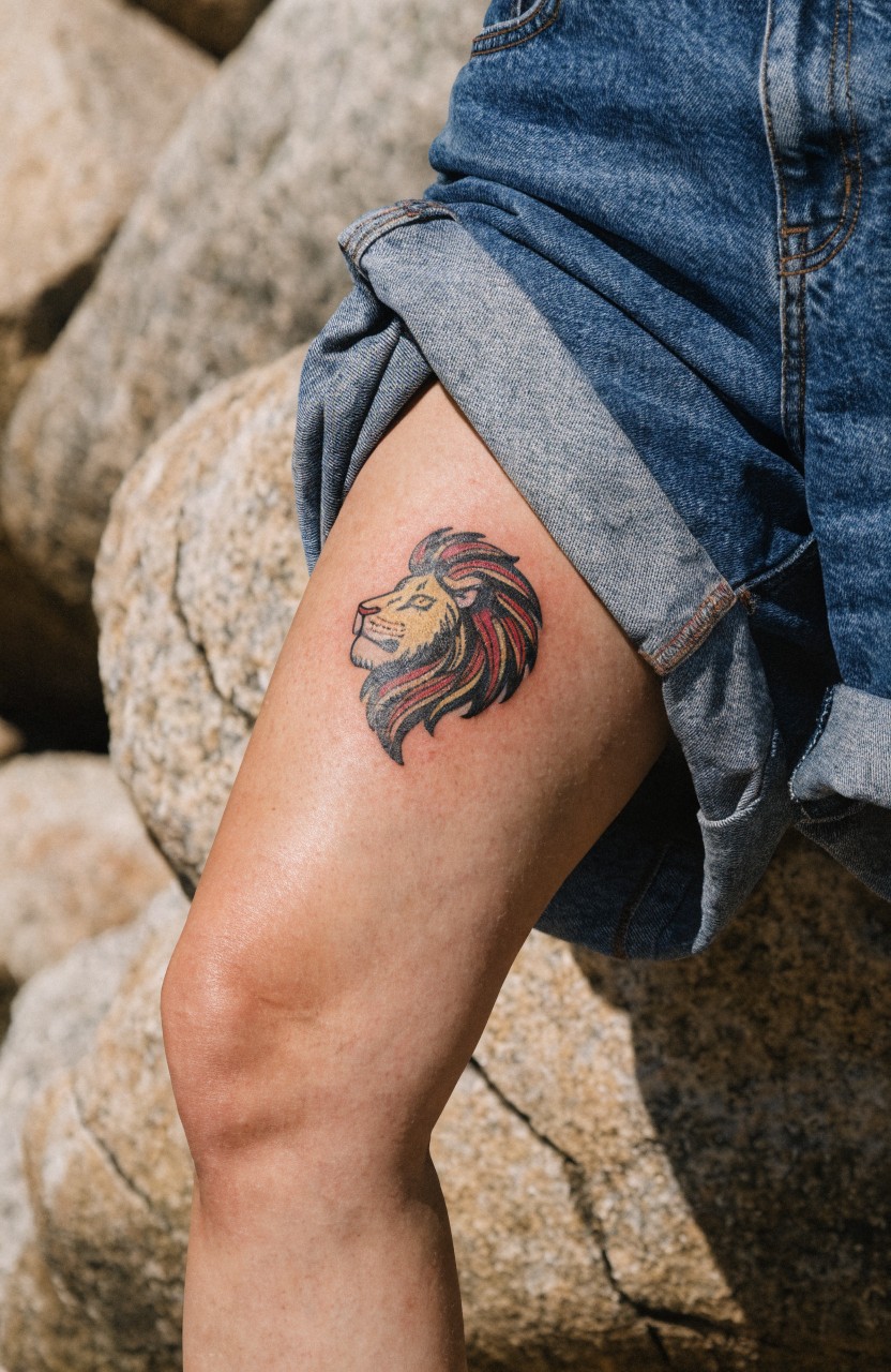 Vibrant lion head tattoo on a woman's outer thigh with flowing red and orange mane, detailed shading, posed among rocks with rolled-up jeans