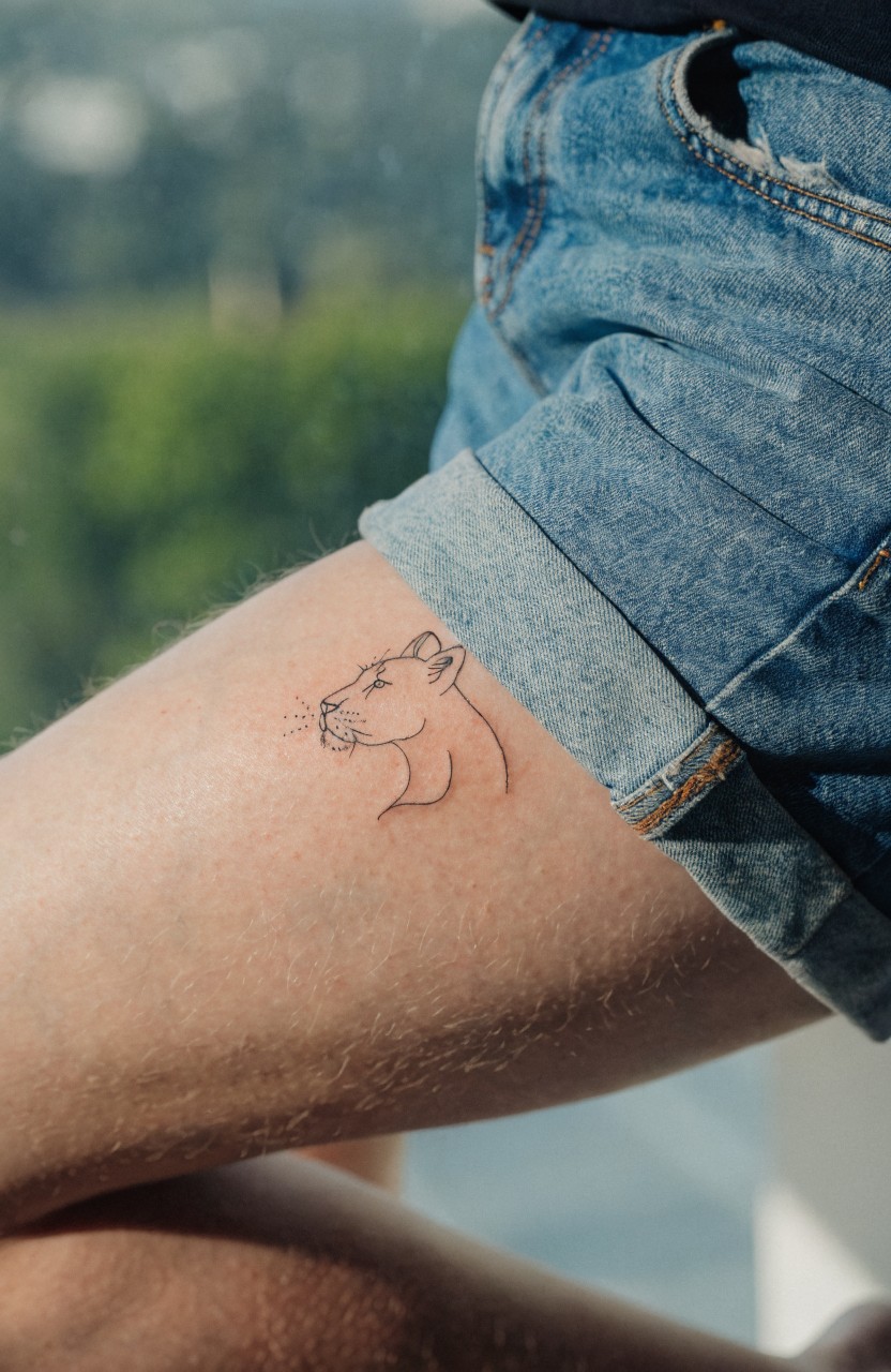 Black line art tattoo of a lioness head in profile on a woman's upper thigh, next to rolled denim shorts