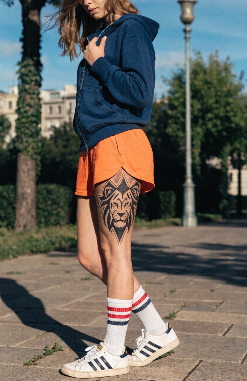 Woman modeling a large realistic black ink lion head tattoo on her upper thigh, detailed with shaded mane and fierce expression, paired with orange shorts, blue hoodie, and white sneakers on a sunny street