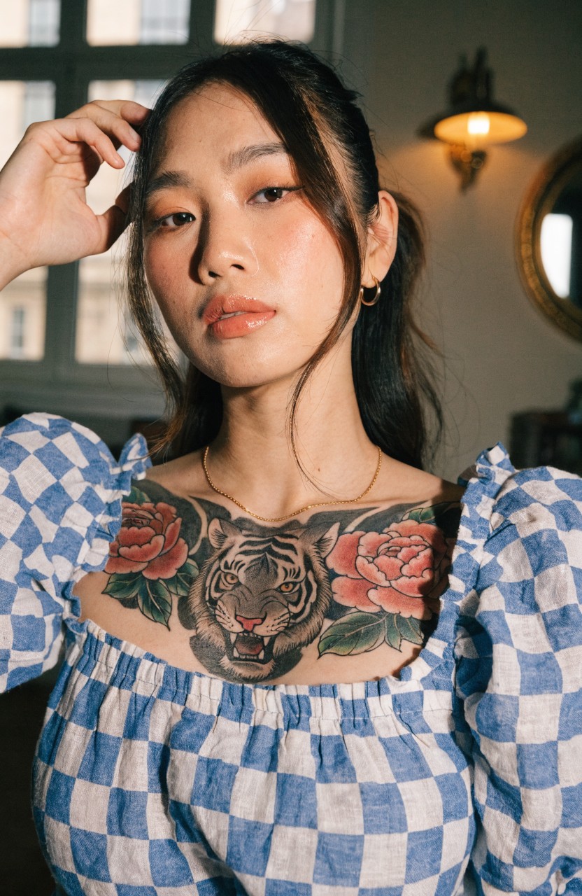 Large traditional Japanese tiger head tattoo with red roses across a woman's upper chest, modeled in a blue checkered off-shoulder top
